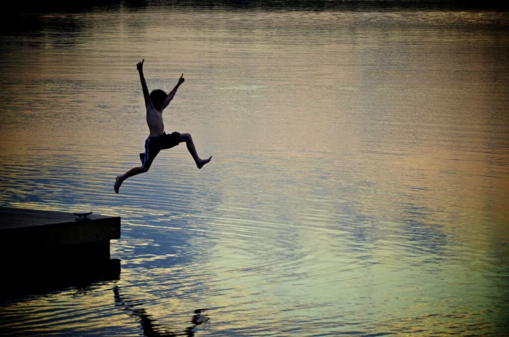 An unidentifiable boy joyfully leaps into a lake at sunset.